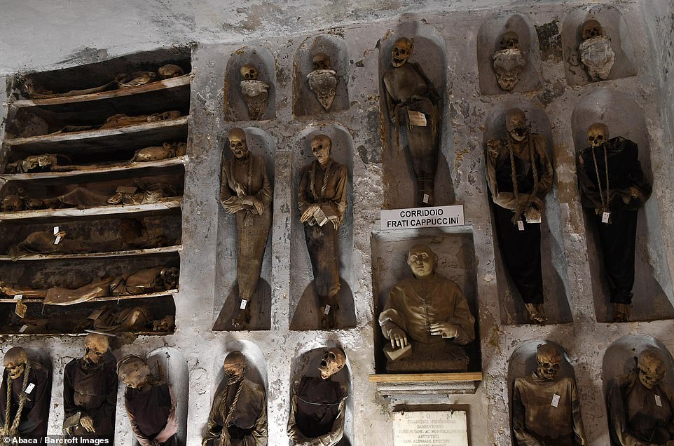 This pictures shows 19 mummies with four separate skulls being stacked on small alcoves at the very top of the wall. Some appear to be wearing ropes around their necks with tags on them