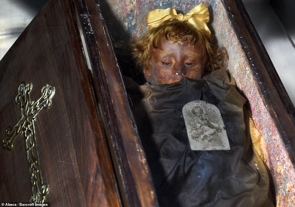 This disturbing pH๏τo shows a small child in a coffin with a silver cross emblazoned on the top. The child still has hair and it wearing a golden bow on top of her head